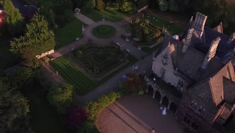 Toma-Aérea-De-La-Mansión-Junto-Al-Lago-Cheltenham-Gloucestershire-Novia-Caminando-A-La-Casa-A-Través-Del-Patio-Y-Los-Jardines-En-La-Hermosa-Luz-Dorada-Del-Atardecer