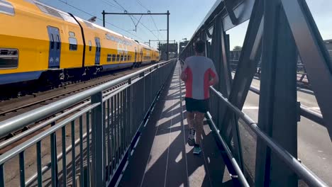 Pasando-El-Tren-Holandés-Siguiendo-El-Rastro-Masculino-Runner-Visto-Desde-Atrás-En-El-Puente-Levadizo-De-Acero-A-Lo-Largo-De-Las-Vías-Del-Tren.