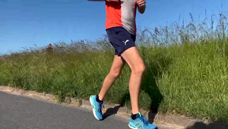 Closeup-of-legs-following-a-middle-aged-male-runner-on-floodplains-valley-dyke-asphalt-with-low-vegetation-along-the-path