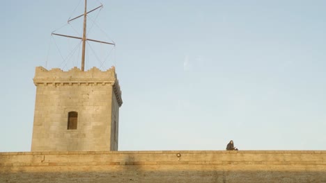 Young-woman-reading-book-during-sunset-on-ledge-of-Montjuic-castle-wall-in-Barcelona-Spain-with-clear-blue-skies