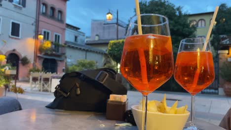 Vista-Estática-Desde-La-Mesa-De-Tres-Amigos-Bebiendo-El-Típico-Aperol-Spritz-Italiano-Al-Atardecer-En-La-Calle-Del-Pueblo-Tradicional