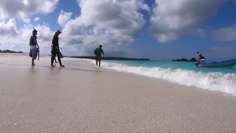 Tourists-with-snorkeling-equipment-are-walking-on-a-white-sand-and-turquoise-water-beach-of-the-Galapagos-islands-as-a-boat-comes-closer-to-the-shore-to-pick-them-up