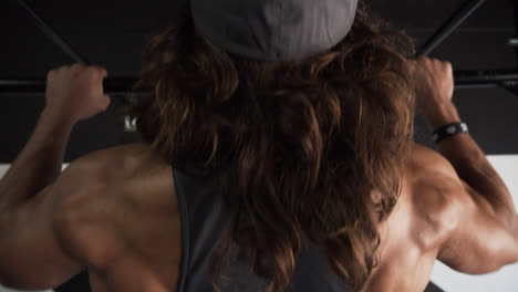 Muscular-man-with-long-hair-doing-pull-ups-in-a-dark-gym-and-showing-his-back-muscles