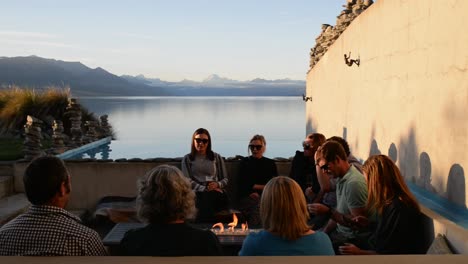 Amigos-Disfrutando-De-Bebidas-Alrededor-Del-Fuego-Al-Atardecer-En-La-Terraza-Con-El-Monte-Cook-Y-El-Lago-Pukaki-Al-Fondo.