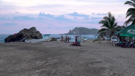 Family-near-rocks-at-beach-waiting-to-observe-the-sunset,-Indio-beer-branded-canopy-right,-Wide-handheld-shot