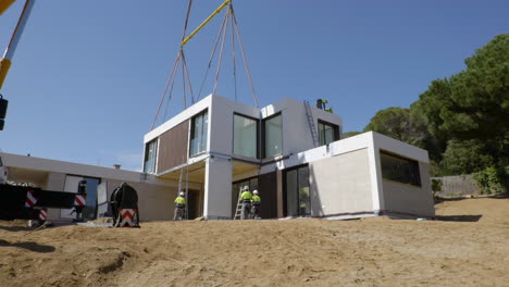 Trabajadores-De-La-Construcción-Instalando-Una-Casa-Modular-Inteligente-Suspendida-Con-Grúa-Pesada-En-El-Sitio-De-Construcción