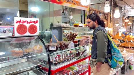 Un-Hombre-Compra-Una-Caja-De-Galletas-En-Una-Pastelería-Local-En-Un-Mercado-De-Agricultores-En-Irán-Esperando-Un-Pastel-De-Galletas-Dulces-Y-Recién-Horneadas-Que-Le-Pagan-A-Una-Comerciante-Que-Trabaja-En-La-Tienda-Con-Productos-Tradicionales