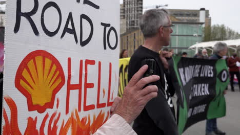 Ein-Aktivist-Für-Den-Klimawandel-Hält-Ein-Plakat-Mit-Der-Aufschrift-„Weg-Zur-Hölle“-Neben-Anderen-Demonstranten,-Die-Vor-Der-Shell-Jahreshauptversammlung-Im-Excel-Ausstellungszentrum-Ein-Transparent-Hochhalten