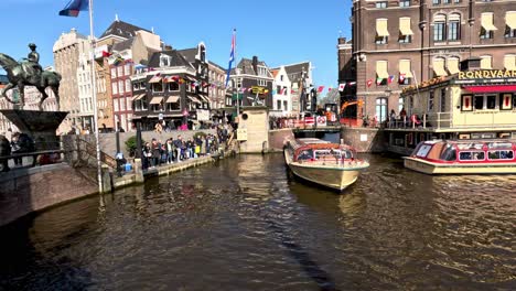 A-cruise-boat-navigating-the-narrow-canal-from-the-corner-of-Langebrugsteeg-alley-and-Rokin-street,-on-the-left-the-sculpture-of-The-equestrian-statue-of-Queen-Wilhelmina-in-Amsterdam,-Netherlands