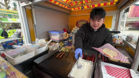 Famoso-Hot-Dog-Estilo-Seattle-De-Un-Vendedor-Masculino-Asiático-En-Un-Carrito-De-Comida-Preparándose-A-La-Parrilla-Con-Cebollas,-Pimientos-Y-Queso-Crema.