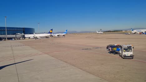 Pequeño-Camión-Cargado-Con-Equipaje-En-La-Pista-Del-Aeropuerto-Rodeado-De-Aviones-En-Madrid,-España