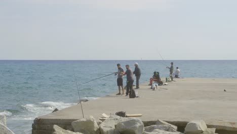 Fishermen-on-stone-pier-in-Barcelona-fishing-for-fish-on-sunny-day-with-slight-wind-and-rough-waters-of-ocean