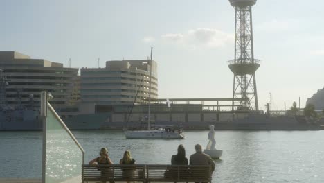 Big-sailboat-setting-sail-out-of-commercial-harbour-in-Barcelona,-people-relaxing-on-benches-at-La-Rambla-del-Mar