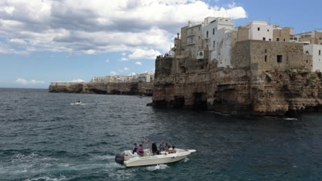 Tourist-boat-tour-near-rocky-coast-with-old-buildings,-Polignano,-Puglia,-Italy