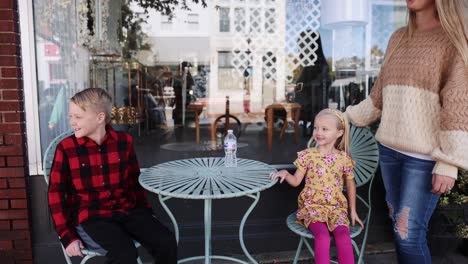 Niño-Y-Niña-Sentados-A-La-Mesa-En-Un-Pequeño-Pueblo-Sonriendo-Al-Aire-Libre