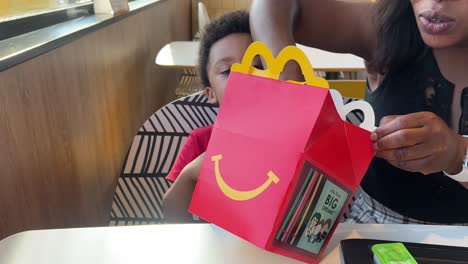 Überraschter-Dreijähriger-Schwarzer-Junge,-Der-Ein-MC-Donald&#39;s-Happy-Meal-öffnet-Und-Neben-Seiner-Schönen-Jungen-Schwarzen-Mutter-Sitzt