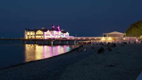 Timelapse-Nocturno-Del-Muelle-De-Sellin-Iluminado-Con-Iluminación-Colorida-Durante-El-Concierto,-Silueta-De-Gente-Caminando-En-La-Playa-De-Guijarros-En-La-Isla-De-Ruegen-Junto-Al-Mar-Báltico