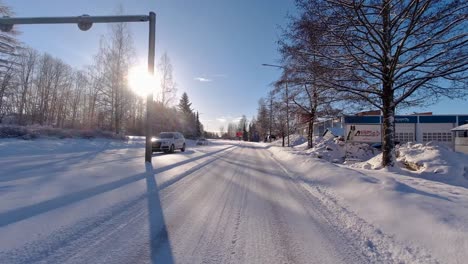 In-winter,-the-car-gracefully-navigates-the-road,-bordered-by-trees,-houses,-and-other-vehicles,-while-the-sun-illuminates-the-surroundings