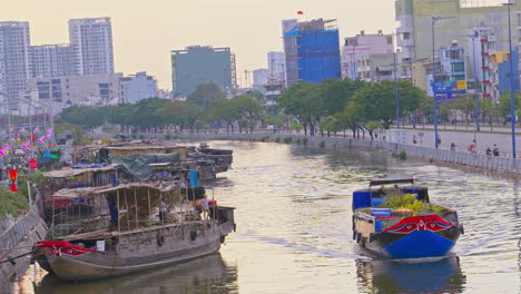 A-boat-carrying-flowers-on-a-city-river,-with-other-boats-docked-at-the-harbor-during-springtime