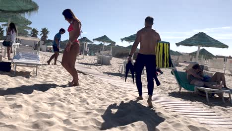 As-a-surfer-walks-past,-a-girl-in-a-bikini-strolls-along-the-wooden-boardwalk-toward-the-beach,-capturing-a-moment-of-coastal-interaction-and-anticipation