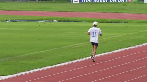 Se-Ve-A-Un-Hombre-Corriendo-En-Una-Pista-Con-Una-Camiseta-Blanca-Número-Once-Y-Una-Gorra-Mientras-Mira-Su-Reloj.