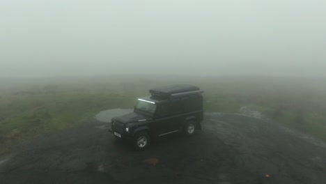 Drone-tilt-up-of-black-Land-Rover-Defender-parked-in-very-cloudy-and-misty-mountain-hills-with-zero-visibility-and-fog-lights-shining