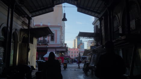 Silhouetted-figures-walk-through-a-shaded-market-street-in-Athens,-Greece,-with-sunlight-peeking-through