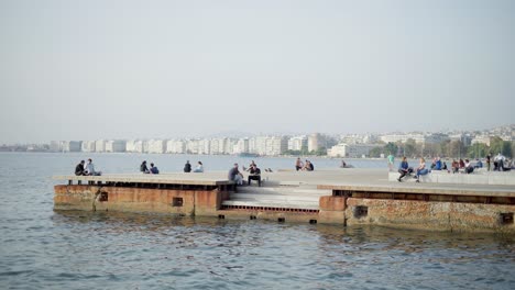 Jóvenes-Sentados-En-El-Muelle-Relajándose-Junto-Al-Mar-En-Tesalónica,-Nuevo-Paseo-Marítimo,-Día-De-Verano.