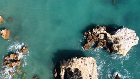 A-revolving-bird's-eye-view-captures-the-rocky-coastline-of-Portugal,-where-the-gentle-splashes-of-sea-waves-caress-the-rugged-formations-in-the-bright-sunshine-of-a-pleasant-day