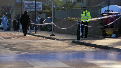 Ein-Stadtpolizist-Bewacht-Den-Eingang-Zu-Einem-Grundstück-Hinter-Einer-Absperrung-Am-Tatort-Eines-Schusswaffenverbrechens,-Während-Zwei-Forensische-Beamte-Die-Hackney-Betreten