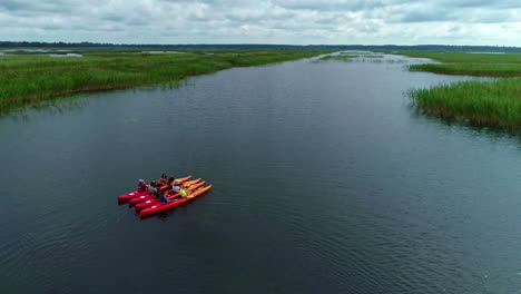 Grupo-De-Personas-Sentadas-En-Kayaks-Esperando-En-Un-Lago-Esperando-Entre-Cañas-Verdes,-Disparo-Aéreo-De-Drones