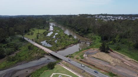 Drone-flying-over-a-green-park-towards-a-river-with-a-bridge-where-cars-passing-by