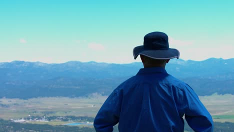 A-white-man-is-on-top-of-the-mountain,-looking-at-the-vast-acres-of-land-and-he-is-pointing-his-fingers-at-something