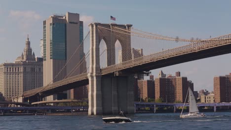 Velero-Y-Lancha-De-Crucero-Por-El-Río-Debajo-Del-Puente-De-Brooklyn-Y-El-Edificio-Verizon-En-El-Fondo---Nueva-York,-Estados-Unidos