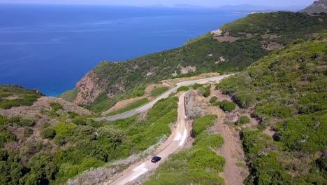 Toma-Aérea-De-Drones,-Siguiendo-Un-Automóvil-Que-Circula-Por-Una-Carretera-Costera-En-Un-Paisaje-Mediterráneo-Con-Impresionantes-Aguas-Azules-Y-Colinas-Verdes,-En-Cerdeña,-Italia,-Al-Sur-De-La-Ciudad-Española-De-Alghero.