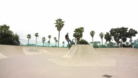 Wide-panning-shot-across-skate-park-as-male-on-BMX-bike-jumps-ramps,-moving-in-closer-as-he-performs-mid-air-trick