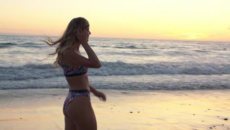 Happy-young-girl-in-bikini-smiles-at-camera-as-she-walks-slow-motions-across-sandy-beach-waves-lapping-into-coast