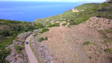 Toma-Aérea-De-Drones,-Siguiendo-Un-Automóvil-En-La-Carretera-Costera-En-Un-Paisaje-Mediterráneo-Con-Impresionantes-Aguas-Azules-Y-Colinas-Verdes,-En-Cerdeña,-Italia,-Al-Sur-De-La-Ciudad-Española-De-Alghero.