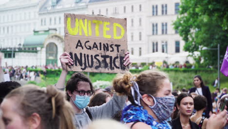 The-words-united-against-injustice-are-written-on-a-paper-banner-which-is-being-held-by-a-young-man-while-protesting-against-violence-and-racism-during-the-corona-pandemic