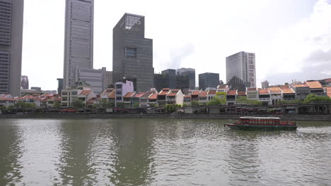 A-full-view-of-the-Singapore-river-facing-Boat-Quay-riverside-pubs,-bars-and-eatery-with-tourist-boats-cruising-by-in-the-afternoon-blue-sky