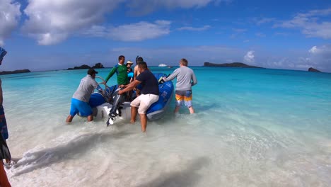 En-Una-Playa-De-Arena-Blanca-Y-Agua-Turquesa-De-Las-Islas-Galápagos,-Varios-Turistas-Suben-Al-Barco-Mientras-Dos-Hombres-Sostienen-El-Barco-Con-Firmeza.