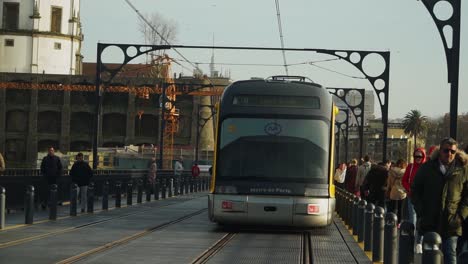 Full-Shot,-Metro-Tram-Train-Leaving-Don-Luis-I-Bridge-in-Porto,-Portugal,-People-Walking-and-Passing-By-in-the-Background