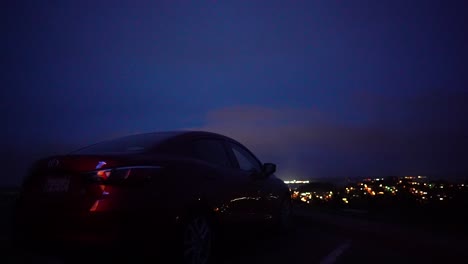 Coche-Estacionado-En-La-Cima-De-Una-Colina-Con-Vistas-A-La-Ciudad-Por-La-Noche