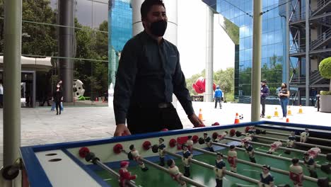 Colegas-De-Oficina-Con-Máscaras-Jugando-Futbolín-Durante-Un-Descanso-Laboral