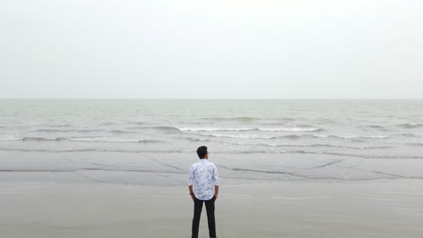 Vista-Aérea-Sobre-Las-Olas-De-La-Playa-Del-Mar-De-Kuakata-Con-Un-Lento-Disparo-Descendente-Para-Revelar-A-Un-Hombre-Solitario-Mirando-Al-Mar