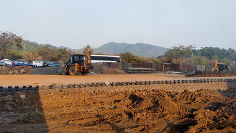 Amidst-the-vibrant-chaos-of-India's-busy-road-traffic,-a-JCB-lifter-diligently-transports-construction-aggregate,-mud,-and-sand-near-the-roadside-|-road-construction,-Indian-road,-traffic,-JCB-machine