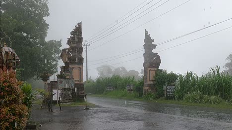 Balinese-local-person-wearing-rain-Poncho-driving-scooter-through-Candi-Bentar-split-Gateway-in-rainy-season,-Bali
