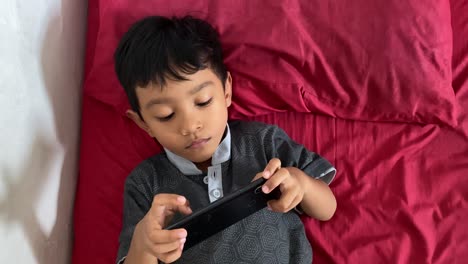 Un-Niño-Asiático-De-4-Años-Jugando-En-Su-Teléfono-Inteligente-Mientras-Está-Acostado-En-La-Cama