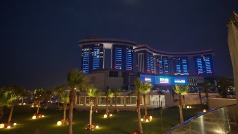 A-view-of-a-large-building-in-El-Alamein,-Egypt-at-night-in-the-middle-of-a-garden-full-of-palms-wide-shot