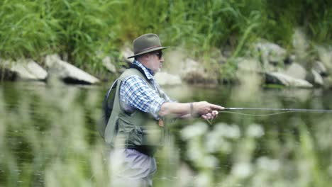 Mann-Beim-Fliegenfischen-In-Einem-Fluss,-Mit-Hut-Und-Sonnenbrille,-Köder-Auswerfend,-Nahaufnahme-Mit-Gras-Im-Vordergrund